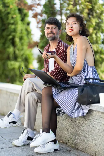 Dışarıda oturan iki farklı arkadaş, parkta bir dizüstü bilgisayarla fikir alışverişinde bulunurken, arkadaşlık ve işbirliğini ifade ediyorlar..