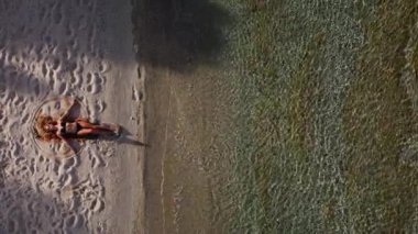 Kumlu kumsalda uzanmış, okyanus kenarında kar meleği şeklinde çekici bir kadın görüntüsü. Yaz tatili ve seyahat konsepti.