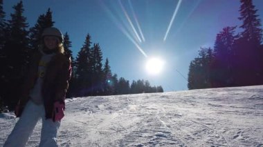 Bir kız snowboardcu güneşli bir yamacın tepesinde duruyor, dalgalar, gülücükler ve tepeden aşağı kayak.