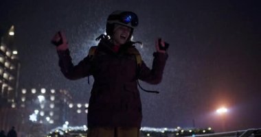 Mutlu genç kadın snowboardcu gece kayak merkezinde yağan karda dans ediyor. Gülümsüyor ve kışın tadını çıkarıyor..