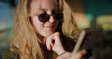 Uzun sarı saçlı ve güneş gözlüklü mutlu genç bir kadının portresi. Telefonu kullanıyor ve gün ışığında dışarıda gülümsüyor..