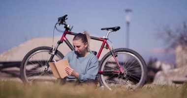Odaklanmış genç bir kadın çimlerin üzerinde kitap okumaktan hoşlanır, bisikleti yakınlarda, açık mavi bir gökyüzünün altında park halindedir..