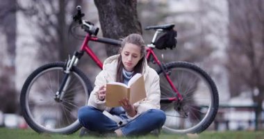 Bir kitaptan fırlamış, kırmızı bisikleti bir ağacın yanında park etmiş, bacak bacak üstüne atmış genç bir kadının huzurlu bir sahnesi..