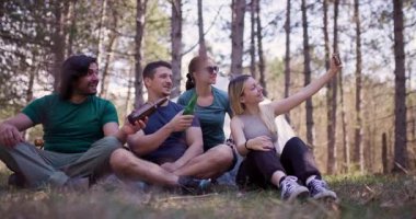 Bir grup arkadaş ormanda oturup selfie çekerken içki içip gülümsüyor. Sıradan açık hava buluşmaları arkadaşlığın ve maceranın özünü yansıtıyor..