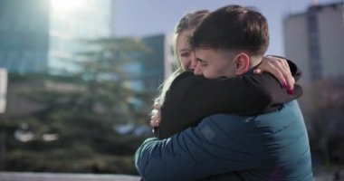 Genç bir çift açık havada sıcak bir kucaklaşmayı paylaşıyor, modern binalarla şehir zeminine neşeyle gülüyorlar. Mutluluk ve yakınlığı gündelik bir ortamda ifade ederler..