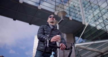 Genç bir adam bisikletiyle açık havada kahve molası veriyor. Modern mimari bir zemin üzerinde. Aktif bir yaşam tarzı ve şehirde boş vakitlerin karışımını gösteriyor..