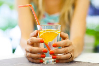 Bir kadının portakal dilimi ve pipetle bardağı tutarken çekilmiş yakın plan çekimleri serinletici bir içkiyi ve açık hava ortamını vurguluyor..