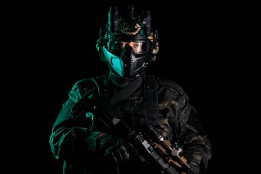 Gece operasyon teçhizatı ve gözlük takan taktik askeri bir şahsiyet, gelişmiş ekipmanları elinde tutarken hazır duruyor. Bu görüntü, gücü, hazırlığı ve profesyonelliği karanlıkta tasvir eder.
