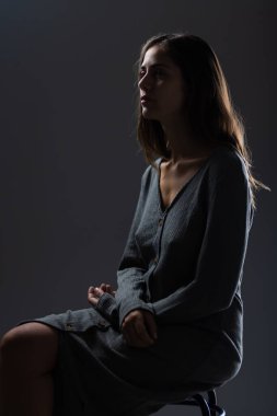 Gri örgü elbiseli genç bir kadının portresi karamsar, düşük ışıklı stüdyo ortamında çekildi. Resim tefekkür ve zarafeti iletir.