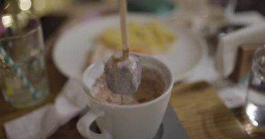 Sıcak çikolata dolu rahatlatıcı bir kupa, rahat bir kafe ortamında karıştırılmış arka planda bulanık tatil ışıkları, sıcak ve davetkar bir atmosfer yaratıyor..