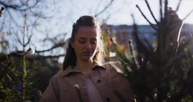 Genç bir kadın, güneşli bir bahçe merkezinde bir çam ağacına dokunuyor ve Noel sezonu boyunca doğa, keşif ve sükunet hissi uyandırıyor..