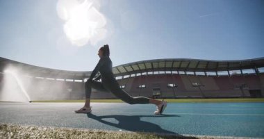 Siluet spor koşucusu kız, sentetik pistte esniyor ve ısınıyor. Açık alan jenerik stadyumunda. Sırtındaki su sulama..