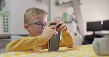 Gözlüklü genç bir çocuk mutfak masasında akıllı bir telefonla oyun oynuyor..