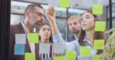 İş arkadaşları proje projelerini tartışıyor. Kafkas iş adamlarından oluşan bir ekip yapışkan notlarla cam duvarın yanında duruyor. Renkli çıkartmalı Scrum metodolojisi. Dikey kamera hareketi.