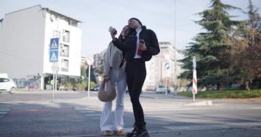 İki kız arkadaş sokakta dikilirken cep telefonlarıyla selfie çekiyor ve sosyal medyayı kontrol ediyor..
