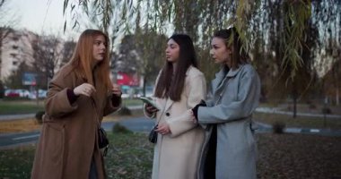 Üç bayan arkadaş kameraya bakıp gülümserken el sallıyorlar. Sonbahar günü. Şehir arkaplanı.