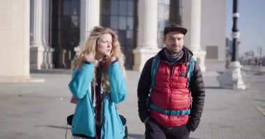 Genç bir adam ve kadın güneşli bir günde sırtlarında sırt çantalarıyla ve kışlık giysileriyle şehir caddesinde yürürler..