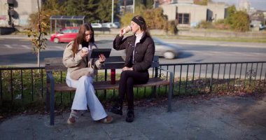 İki bayan arkadaş parkta bir bankta kahve paylaşıyor, birlikte tablet okuyor ve sonbahar havasının tadını çıkarıyorlar..