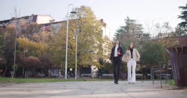 Güneşli bir sonbahar gününde iki bayan arkadaş parkta yürüyor, kahvelerini yudumluyor ve birbirlerine eşlik ediyorlar..