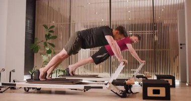 İki birey, çekirdek gücü ve esneklik için çeşitli egzersizler sergileyerek bir pilates oturumuna katılıyorlar. Ayarda parlak ekipmanlar ve minimalist bir tasarım yer alıyor..