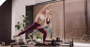 İki kadın, doğal ışık ve yeşillikle dolu şık bir stüdyoda ileri düzey pilates teknikleri uyguluyor. Sahnede modern ekipman ve sakin bir atmosfer yer alıyor..