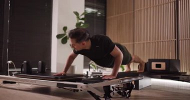 Bir adam, şık bir fitness stüdyosunda bir reformcuya şınav çekerek gücünü gösteriyor. Parlak alanda bitkiler ve modern ekipmanlar var, ilham verici bir çalışma ortamı yaratıyorlar..