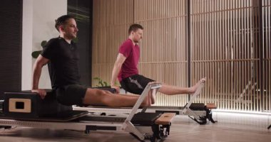 İki adam zarif bir stüdyoda reformcular üzerinde pilates egzersizleri yapıyor. Öğleden sonraki antrenman sırasında hareketlerini sergiledikleri için ortam sakin ve odaklanmış durumda..