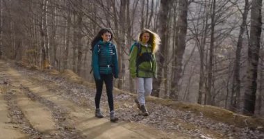 İki bayan arkadaş, sırt çantaları ve yürüyüş botları giyerek dağlarda yürüyüş yapmaktan zevk alıyorlar. Toprak bir yolda yürüyor, konuşuyor ve gülüyorlar..
