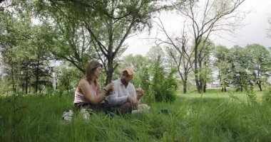 Bir çift, yemyeşil bir parkta, ağaçlarla çevrili bir pikniğin tadını çıkarıyor. Doğanın huzuru içinde yiyecek ve içeceklerinin tadını çıkarıyorlar..