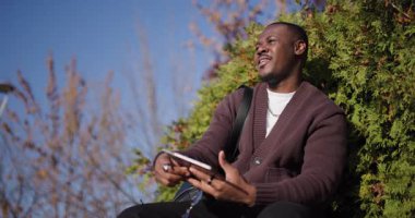Genç siyah bir adam parkta güzel bir sonbahar gününün tadını çıkarıyor, dinleniyor ve tabletini kullanıyor. Cihazıyla etkileşime girerken gülümsüyor ve işaret ediyor..
