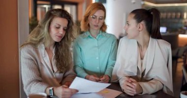 Üç iş kadını, önemli belgeleri inceleyerek ve çağdaş bir kafe ortamında fikirlerini paylaşarak ortak bir tartışmaya giriştiler. Bağlılıkları takım çalışması ve profesyonelliği yansıtıyor..