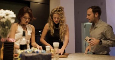 Arkadaşlar modern bir mutfakta yemek hazırlayıp sohbet ediyorlar. Biri telefonunu kullanırken, diğerleri peynir kesip abur cubur hazırlıyor..