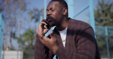 Genç bir adam açık hava basketbol sahasında bir bankta oturuyor, telefonuyla konuşuyor ve elleriyle işaret ediyor..