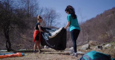 İki genç kadın dağlarda bir nehrin yanında bir çadır kurdular. Kamp yerlerini gece için hazırlıyorlar..