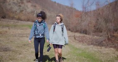 İki bayan arkadaş, güneşli bir günde sohbet ederek ve sırt çantalarını taşıyarak doğa yürüyüşünden zevk alırlar..
