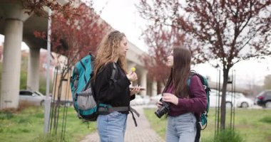 Sırt çantalı ve kameralı iki genç kadın şehir parkında seyahat planlarını tartışıyor..