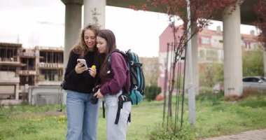 Sırt çantalı ve kameralı iki genç kadın telefona bakıp gülüyorlar. Kaldırımda duruyorlar. Ağaçların önünde ve otoyolun altında..