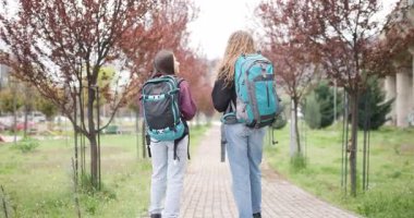 Sırt çantalı iki genç kadın parkta yürüyüp fotoğraf çekerken sohbet ediyorlar..