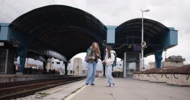 Sırt çantalı ve kameralı iki genç kadın tren istasyonunda yürüyüp konuşuyorlar..