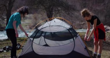 İki bayan arkadaş güneşli bir günde ormanda bir nehrin kenarında çadır kuruyorlar. Kamp gezisindeler..