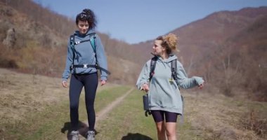 Güneşli bir günde yürüyüş yapan iki bayan arkadaş. Sırt çantaları ve yürüyüş botları giyiyorlar ve manzaranın tadını çıkarıyorlar..