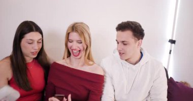 Üç arkadaş birlikte oturup gülüyorlar ve konuşuyorlar. Beyaz bir fotoğraf makinesiyle çekilmiş bir fotoğrafı izliyorlar..