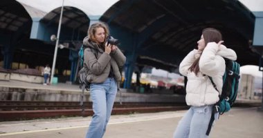 Sırt çantalı iki genç bayan bir tren istasyonundalar. Biri diğerinin fotoğraf makinesiyle fotoğrafını çekiyor. Yolculukları için heyecanlılar..