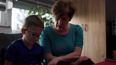 Büyükannem torununa ev ödevinde yardım eder. Çocuk kalemle bir deftere yazıyor ve gözlük takıyor. Kanepede oturuyorlar..