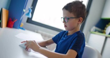 Gözlüklü genç bir çocuk masasında oturuyor ve dikkatlice bir parça kağıt katlıyor, muhtemelen origami yapıyor..