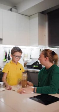 Genç bir çocuk bir bardak süte çikolata tozu dökerek sıcak bir mutfakta annesinin yardımıyla lezzetli bir çikolatalı süt içeceği yaratıyor..