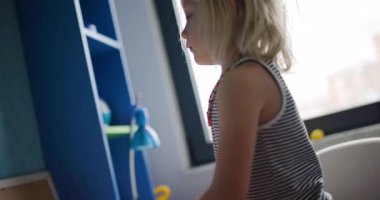 Sevimli küçük bir kız, yatak odasındaki masasında renkli kalemler ve kağıtlarla mutlu bir şekilde oynuyor..