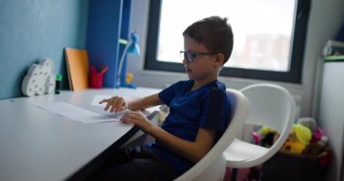 Gözlüklü genç bir çocuk masasında oturur ve bir parça kağıt katlar. Odaklanmış ve işine odaklanmış durumda..