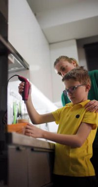 Bir anne, oğlunun sağlıklı bir içecek yapmak için mutfakta el blenderi kullanmasına sevgiyle yardım eder. Birlikte çalışırlar, besleyici bir atıştırmalık oluştururlar ve bir bağ kurma anını paylaşırlar..