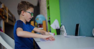 Genç bir çocuk masasında oturuyor, beyaz kağıtları dikkatlice katlıyor. Gözlüklü ve mavi tişörtlü..